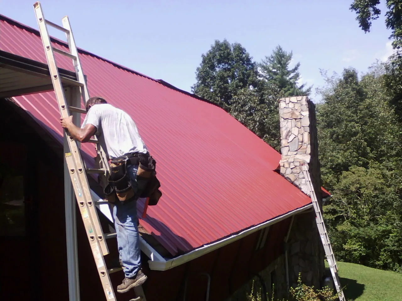 Roofer on a ladder beside a red metal roof and stone chimney — Soffit & Fascia Repair by Roofing Brothers in Playa Del Rey